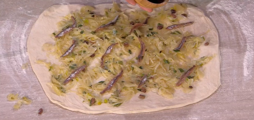 Nel frattempo, in padella, facciamo stufare gli sponsali (o cipollotti) affettati con un filo d'olio e del sale. Lasciamo raffreddare. Stendiamo l'impasto con il mattarello, in modo da ottenere un rettangolo spesso circa mezzo cm. Distribuiamo sopra i cipollotti stufati, i filetti di acciughe sgocciolati ed interi, l'uvetta. Arrotoliamo l'impasto sul ripieno, partendo dal lato lungo. Ottenuto un salsicciotto, lo avvolgiamo su se stesso, a chiocciola. Inseriamo quest'ultima all'interno di una tortiera (da 30 cm) unta d'olio, copriamo e lasciamo lievitare fino al raddoppio. Spennelliamo la superficie con l'olio e cuociamo in forno caldo e statico a 250° per 20 minuti; abbassiamo a 200° e lasciamo cuocere ancora 20 minuti.